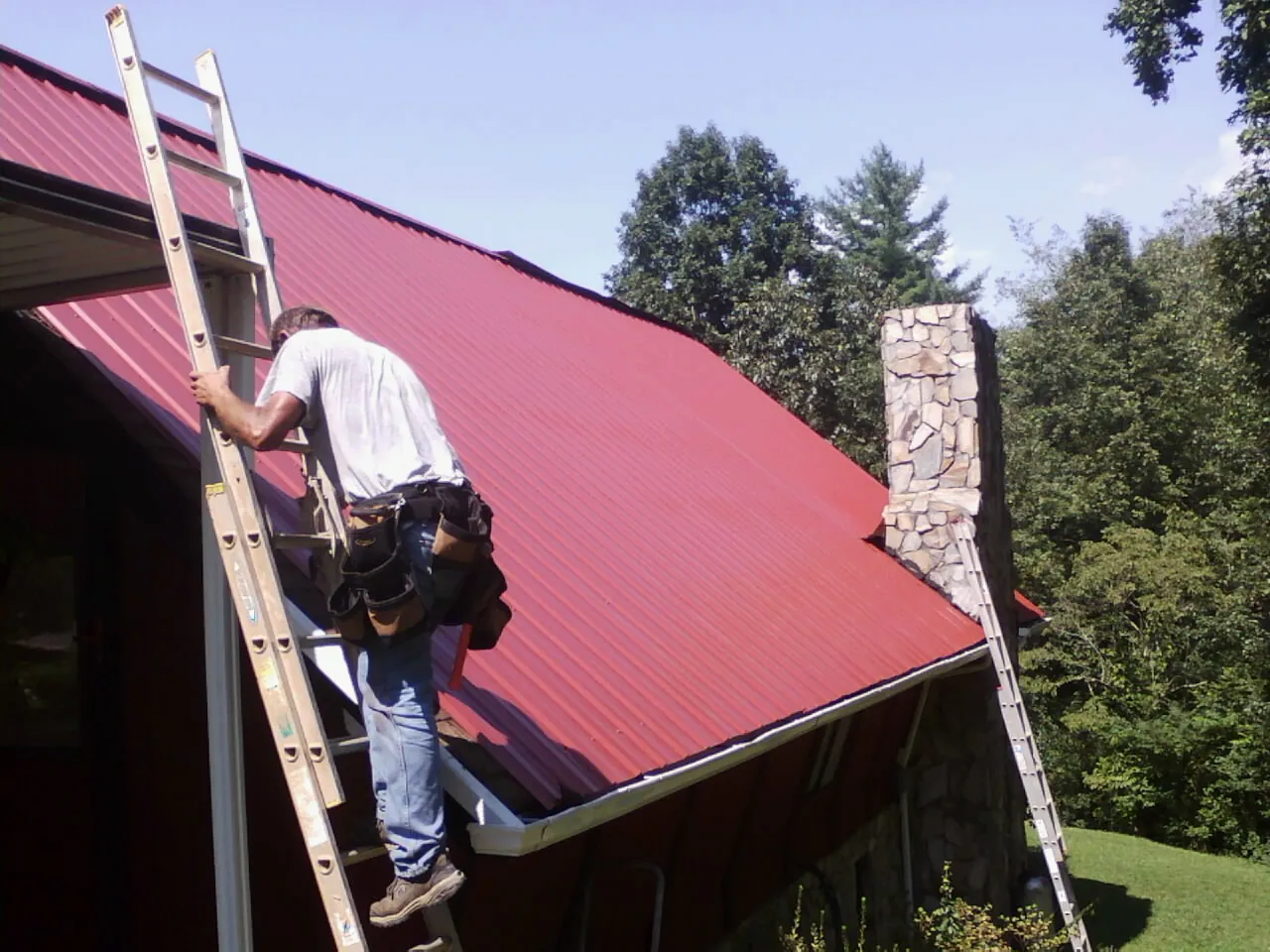 Roofer on a ladder beside a red metal roof and stone chimney — Soffit & Fascia Repair by Roofing Brothers in Johnsonburg